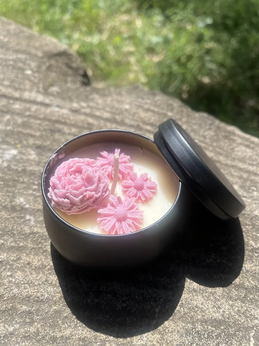 A candle in a black tin topped with pink wax flowers, on a sunny sandstone ledge